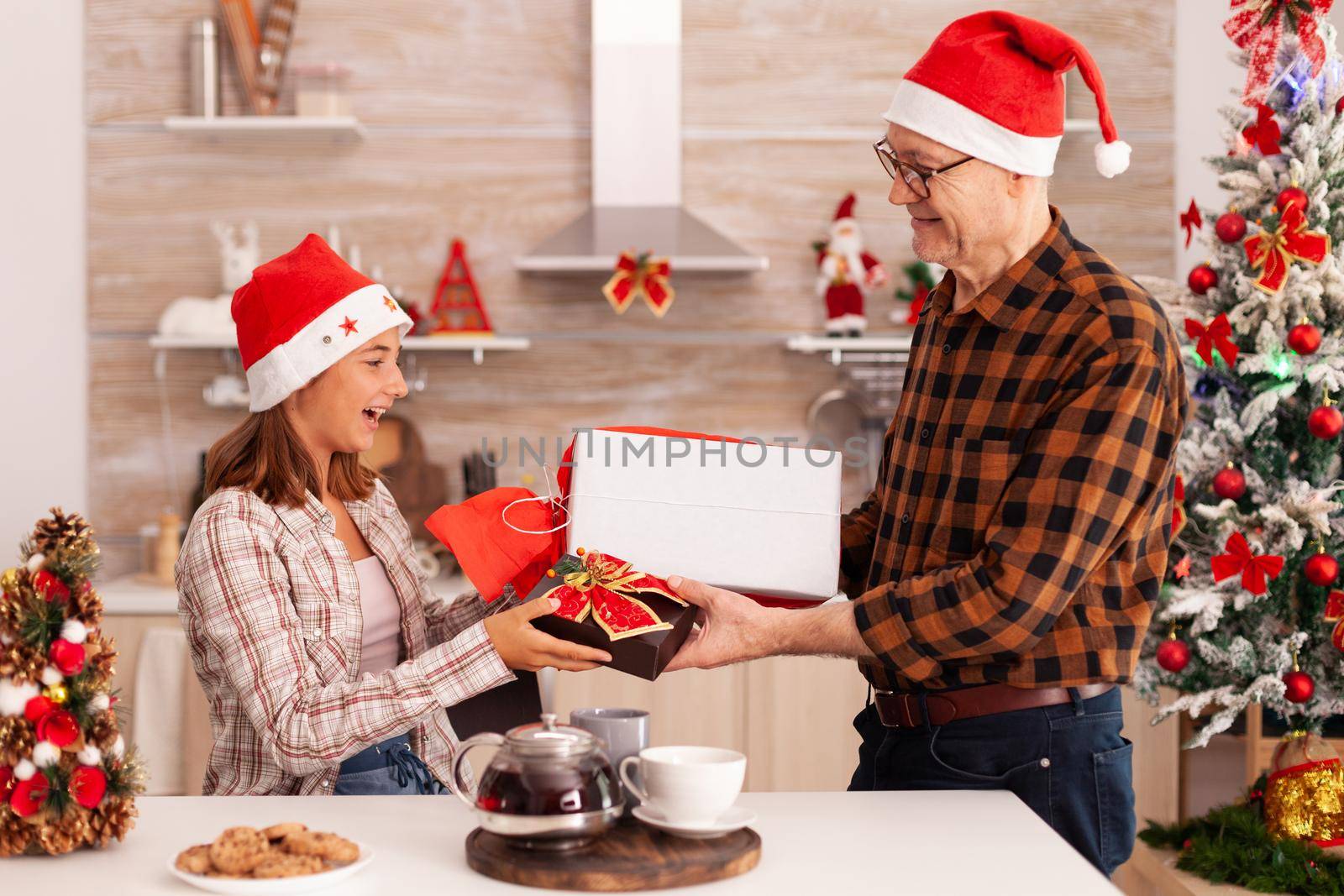 Grandfather surprising granddaughter with xmas wrapper gift enjoying christmas holiday together in decorated kitchen. Child celebrating winter season changing present with ribbon on it with grandpa