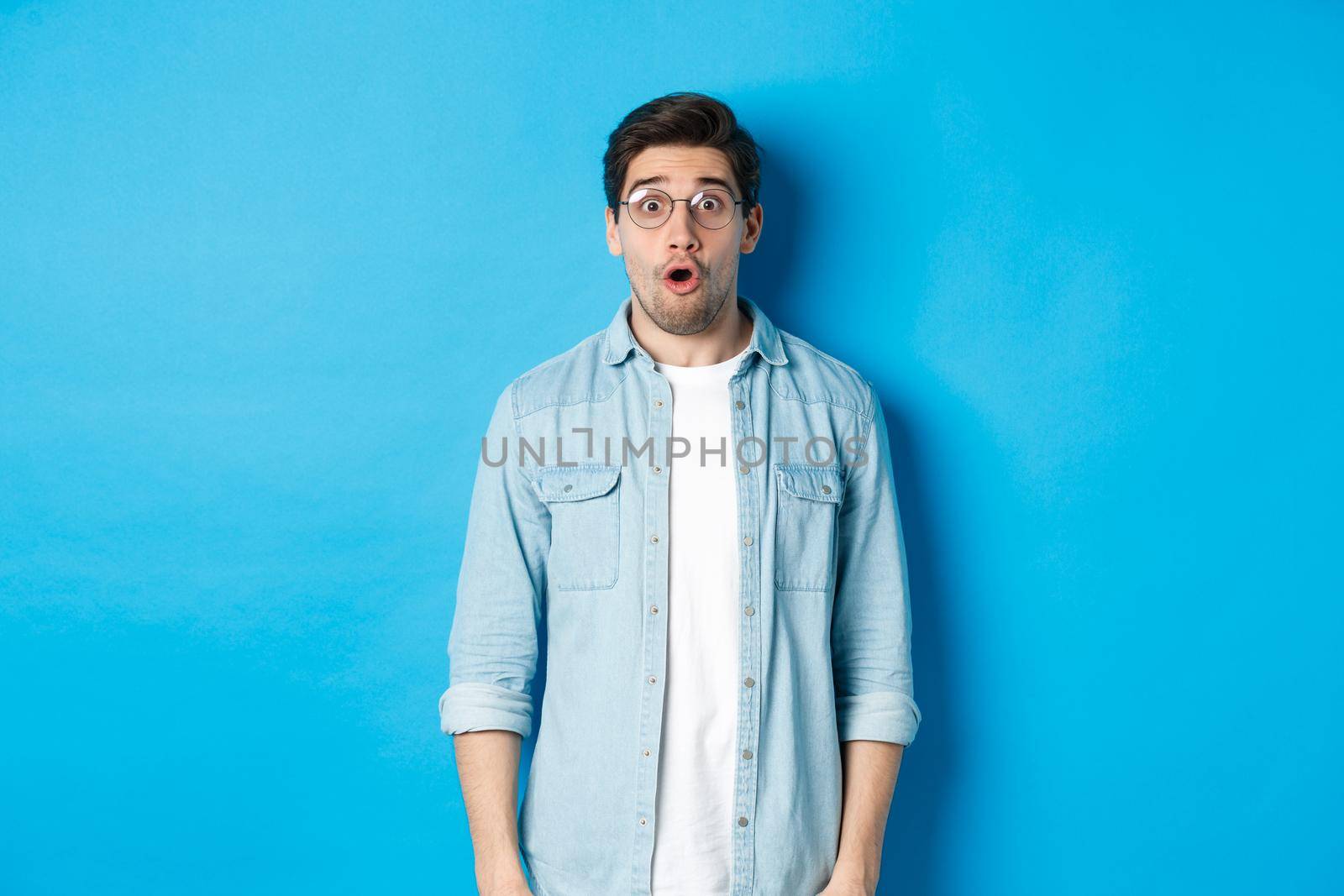 Surprised man in glasses looking at promo offer, saying wow amazed, standing over blue background by Benzoix