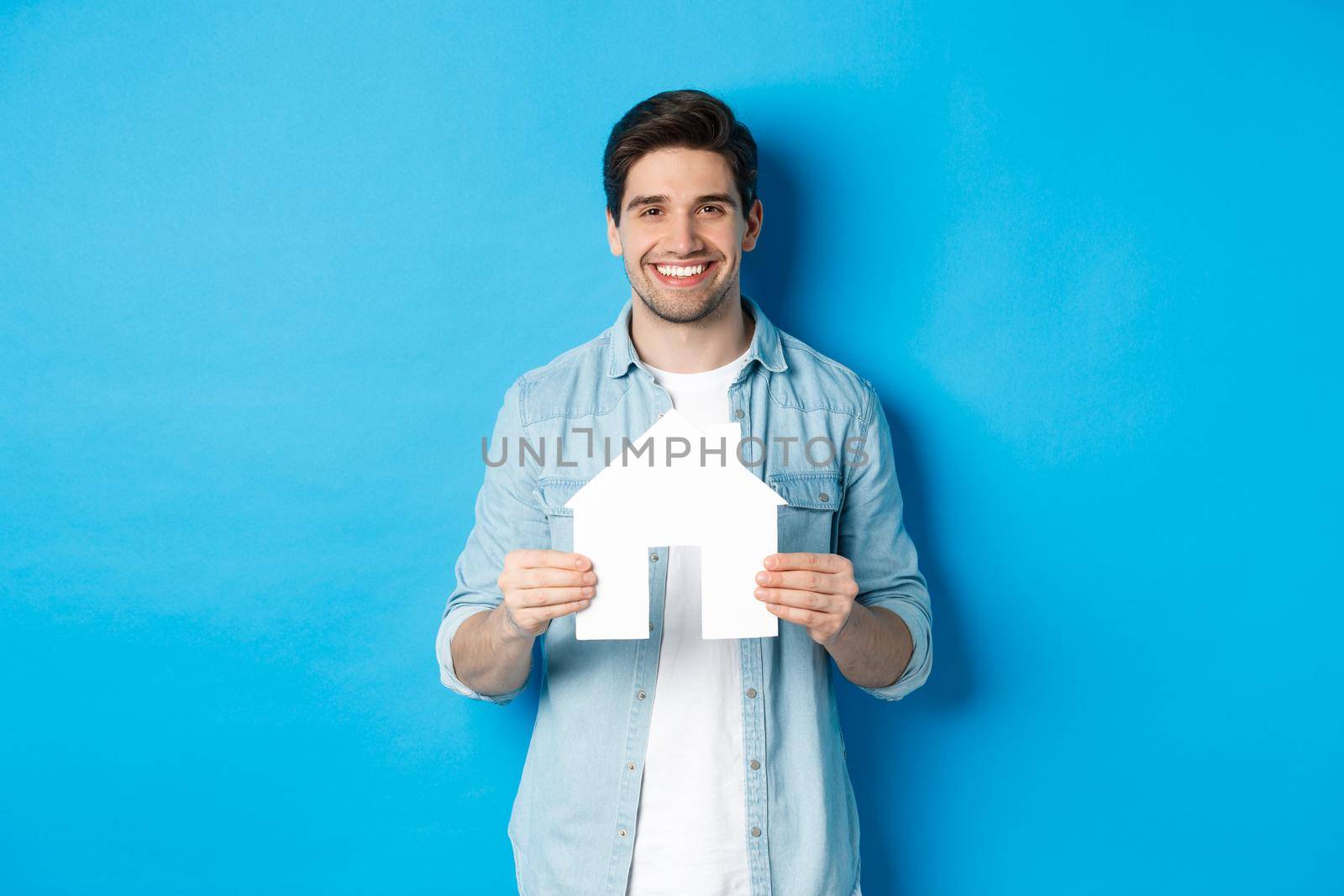 Insurance, mortgage and real estate concept. Smiling young man holding house model, searching apartment for rent, standing against blue background.