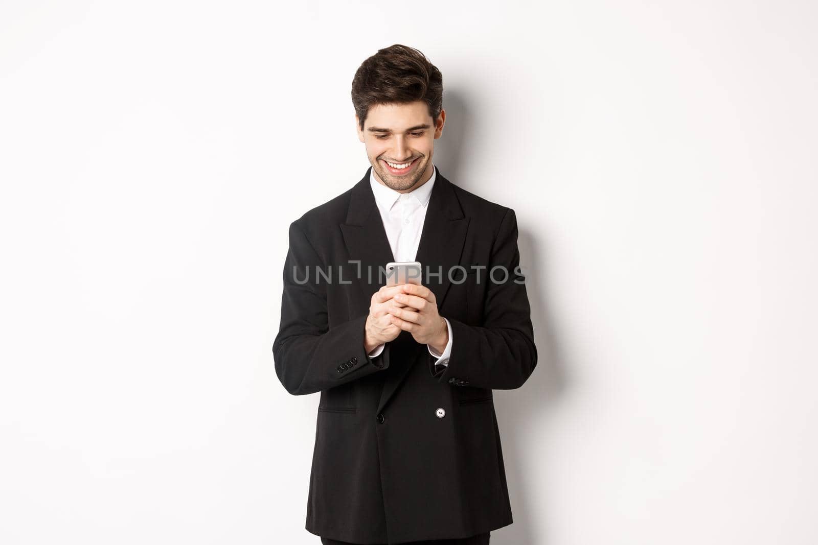Portrait of handsome stylish businessman in black suit, writing a message, smiling and looking at smartphone, standing over white background by Benzoix