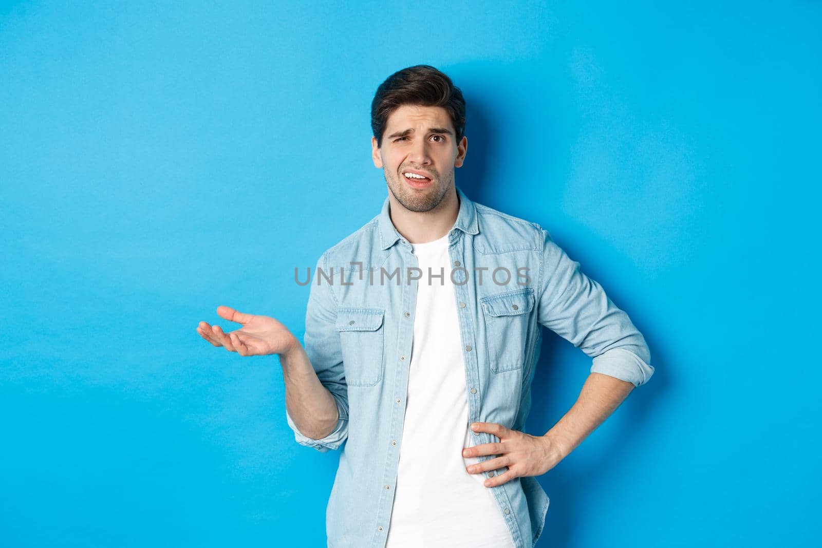 Careless man looking at something disappointing, complaining and judging bad product, standing unamused against blue background.