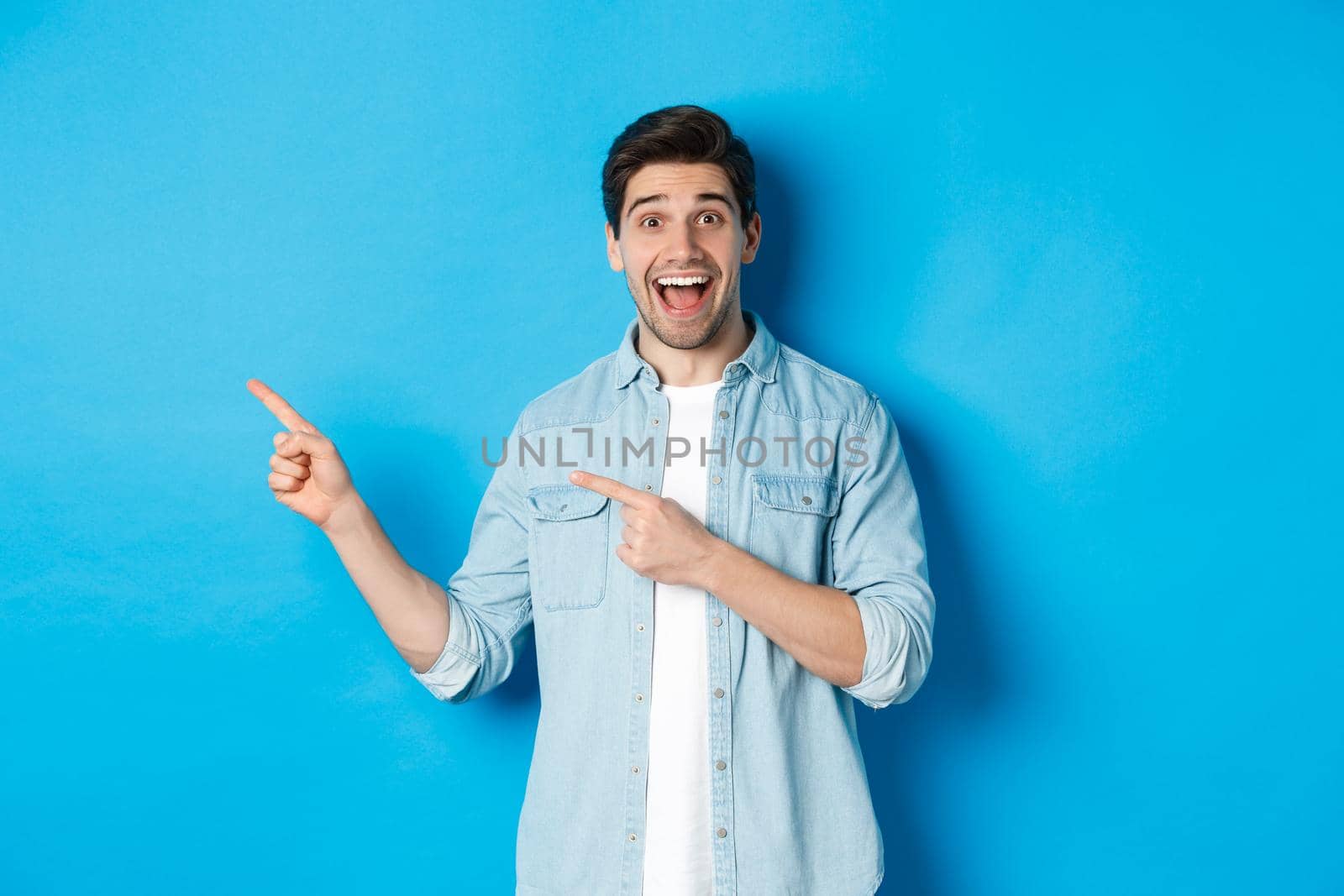 Excited handsome 25s years man with beard, pointing fingers left and smiling amazed, standing against blue background by Benzoix