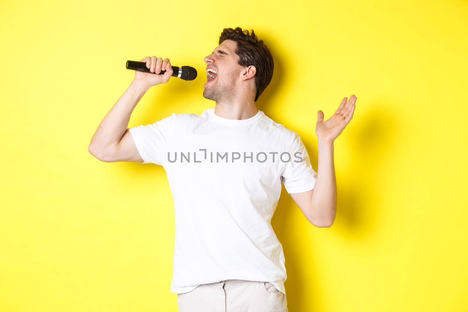 Young man singer holding microphone, reaching high note and singing karaoke, standing over yellow background.