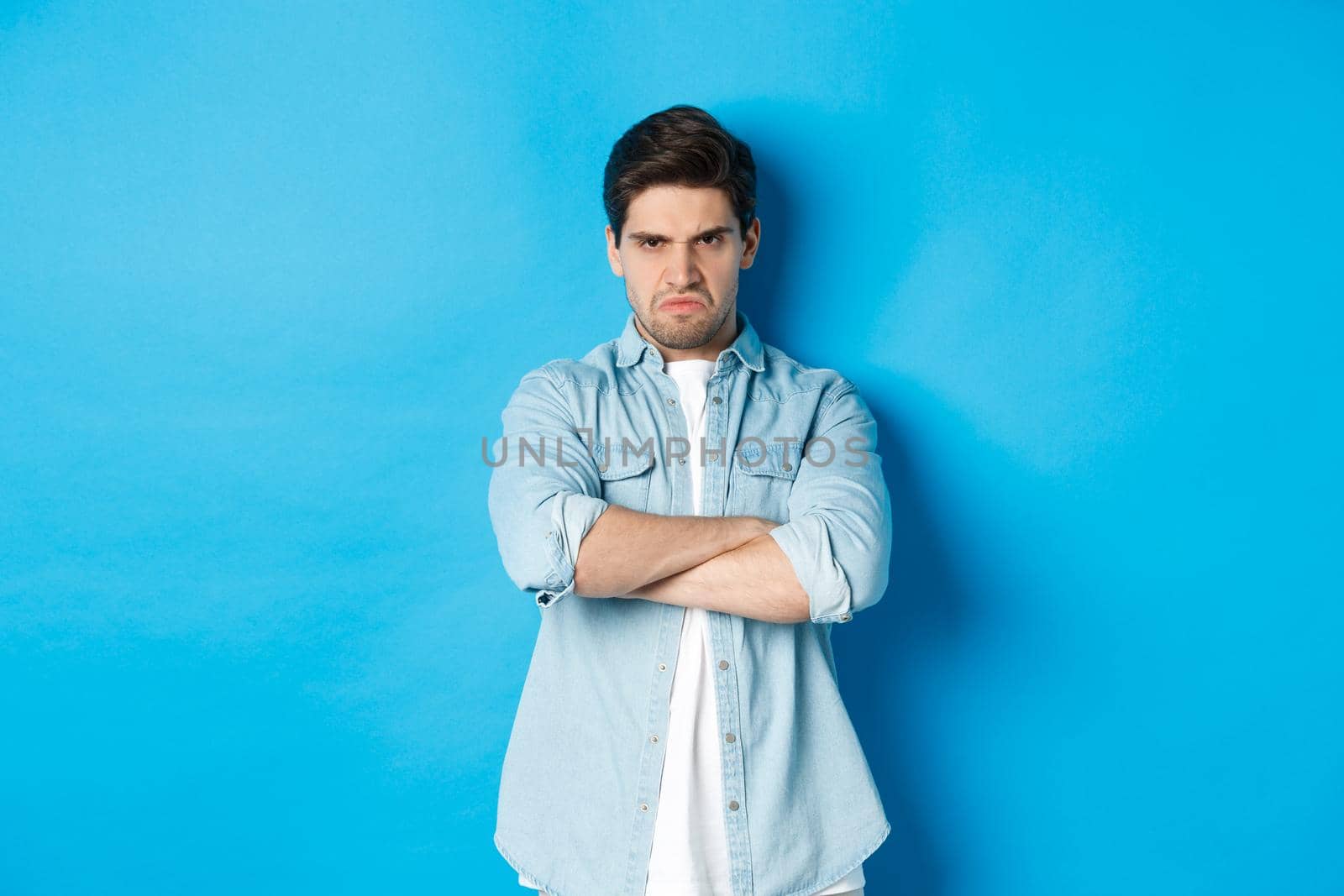 Angry bearded guy crossing arms on chest, frowning, looking mad and offended, standing against blue background.