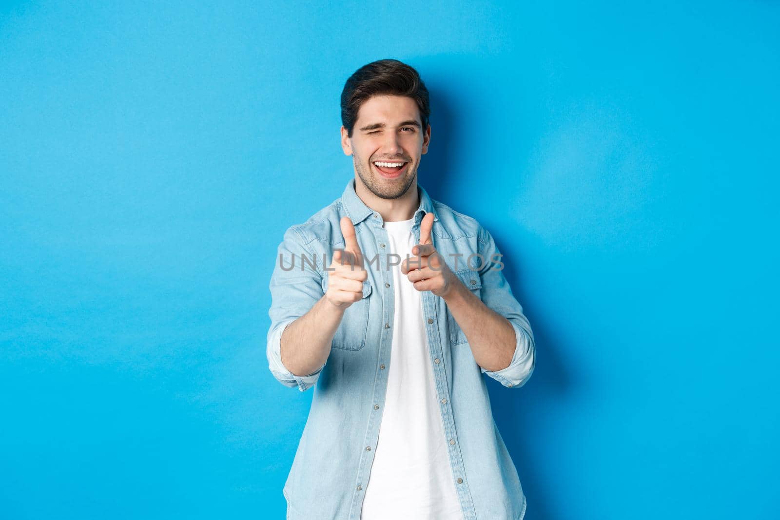 Cheeky handsome guy pointing fingers at you, winking flirty, standing in casual outfit against blue background.