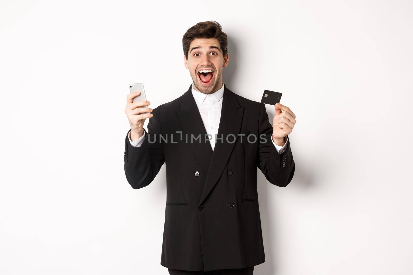 Handsome businessman in black suit smiling, showing credit card and money, looking amazed, standing against white background by Benzoix