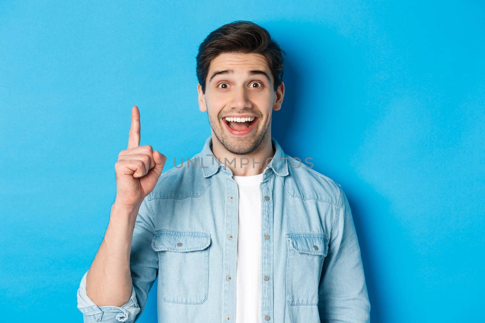 Close-up of handsome bearded guy smiling, showing finger number one, standing over blue background.