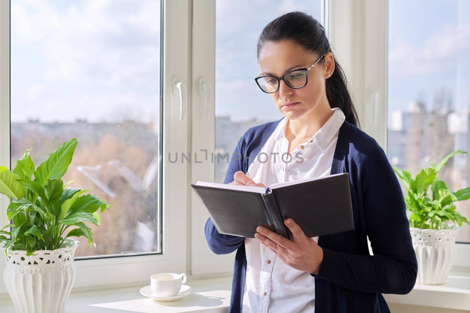 Adult woman with notepad pencil at home near window. Middle-aged female with glasses writing in notebook. Blogger, author, business, psychologist, counselor, teacher, work, hobby, leisure, people 40s