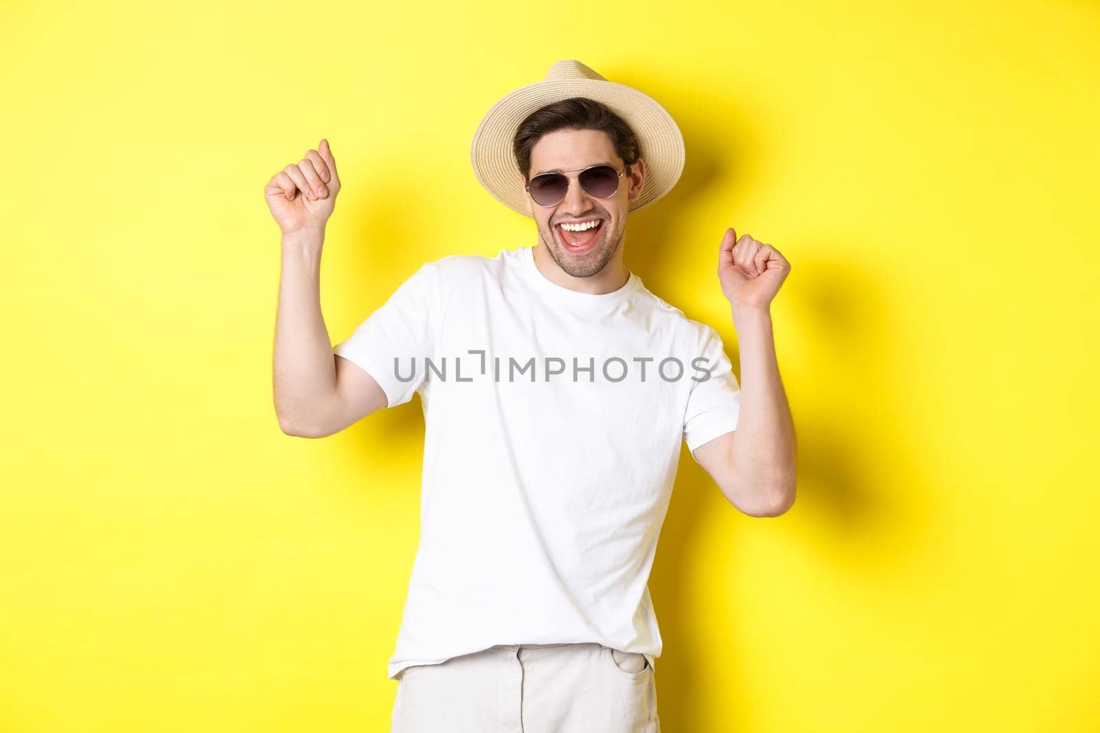 Tourism, travelling and holidays concept. Happy caucasian guy dancing and having fun on vacation, wearing sunglasses with straw hat, standing against yellow background.