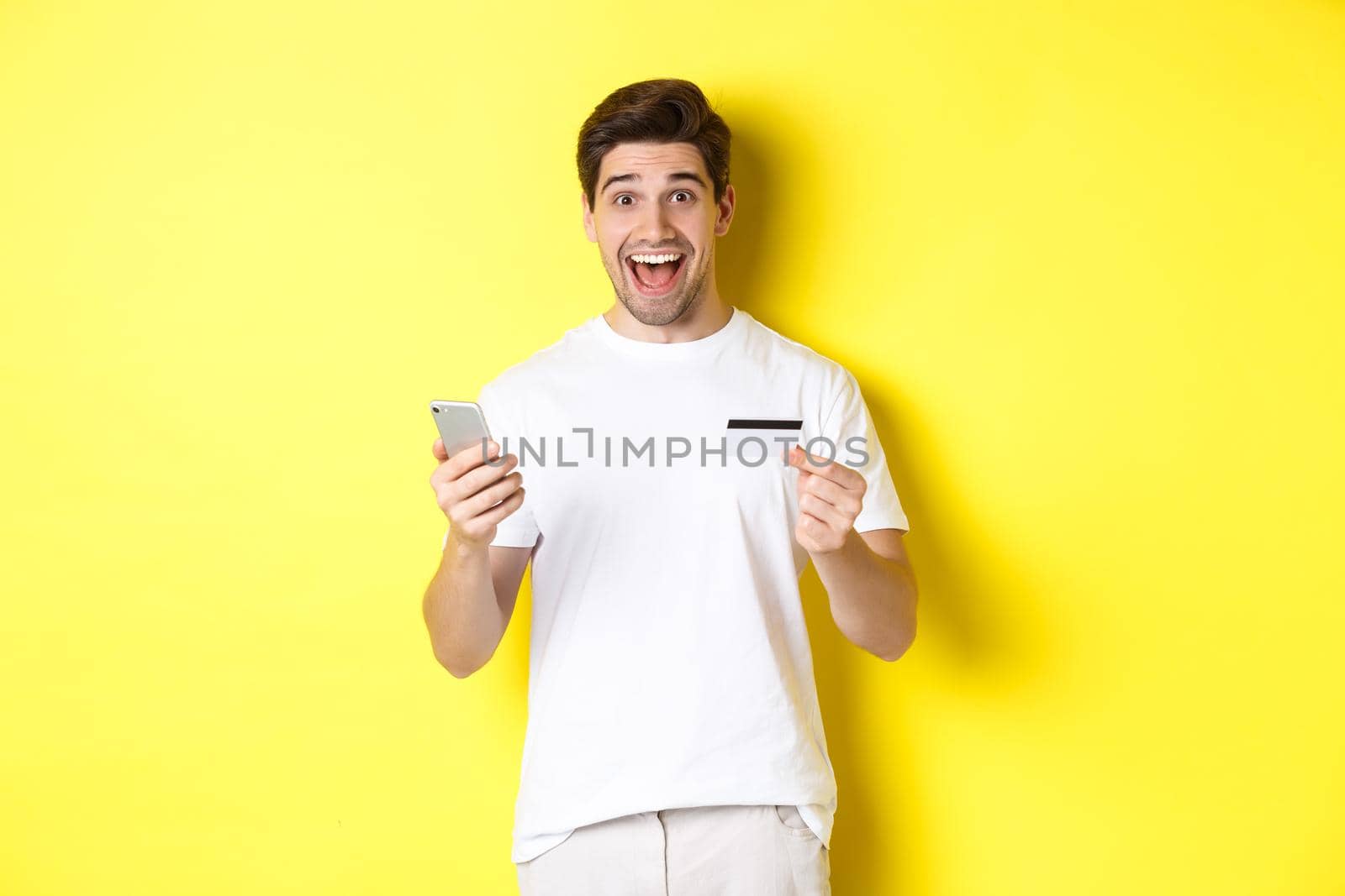 Happy male buyer holding smartphone and credit card, concept of online shopping in internet, standing over yellow background.