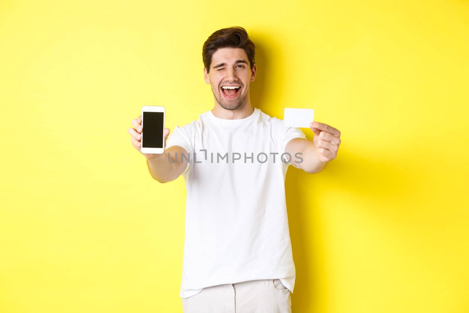 Happy man showing good online offer on mobile phone screen, holding credit card and winking, standing over yellow background.