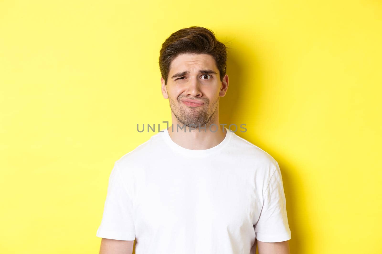 Close-up of dispelased young man in white t-shirt looking doubtful, grimacing unsatisfied, standing over yellow background.