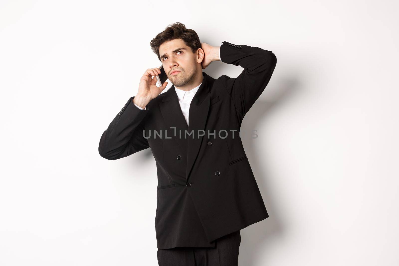 Image of indecisive businessman talking on phone and thinking, looking doubtful, making decision, standing over white background.