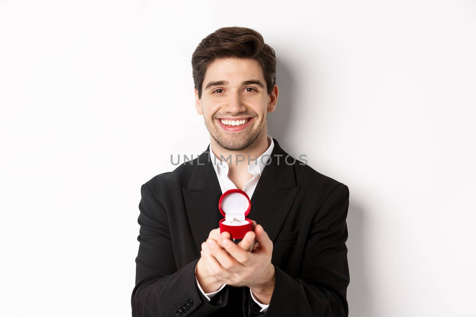 Image of handsome man looking romantic, open small box with engagement ring, making a proposal and smiling, standing against white background.