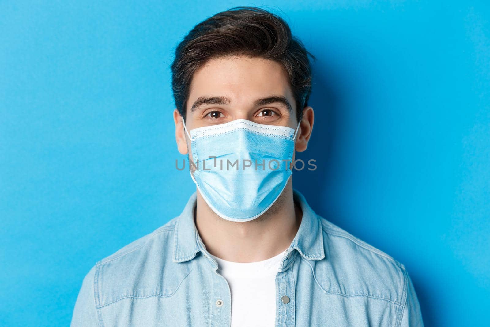 Concept of covid-19, pandemic and quarantine. Close-up of happy guy in medical mask looking at camera, standing over blue background by Benzoix