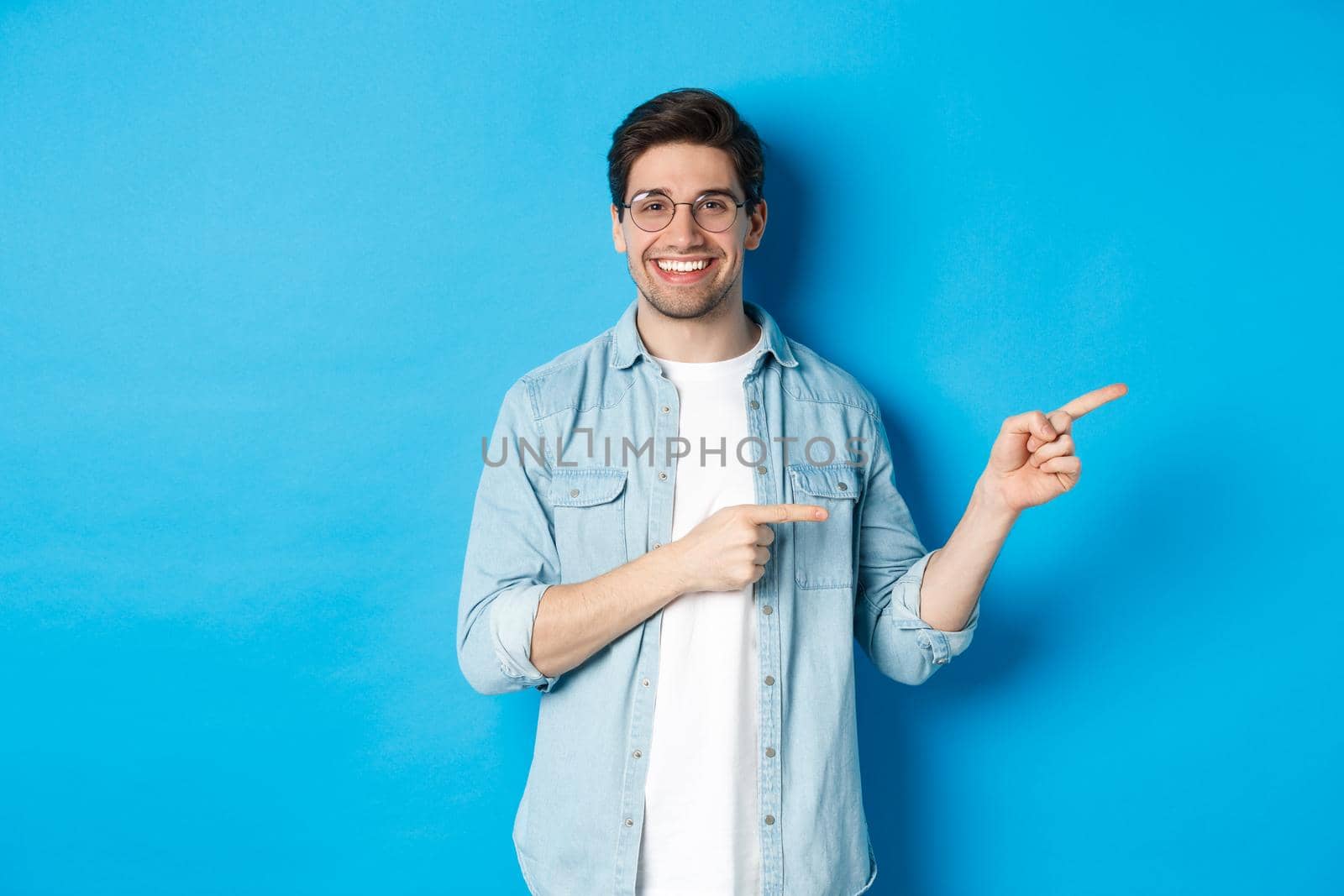 Handsome caucasian male model in glasses, pointing fingers right at your logo, showing copy space, standing over blue background.