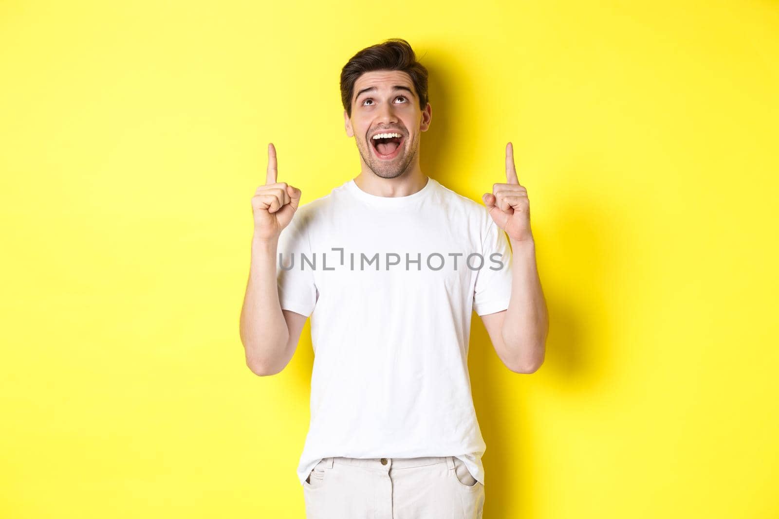 Cheerful young guy in white t-shirt reacting to promo offer, pointing and looking up with amazement, standing over yellow background by Benzoix