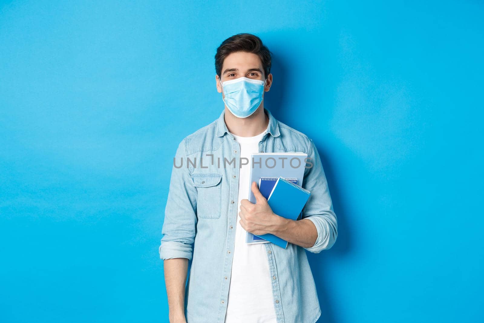 Education, covid-19 and social distancing. Guy student in medical mask looking happy, holding notebooks, standing over blue background.