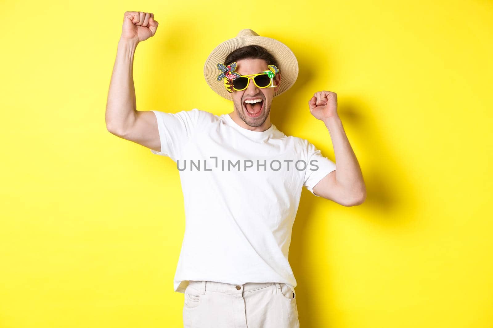 Concept of tourism and lifestyle. Happy lucky guy winning trip, rejoicing and wearing holiday outfit, summer hat and sunglasses, yellow background.