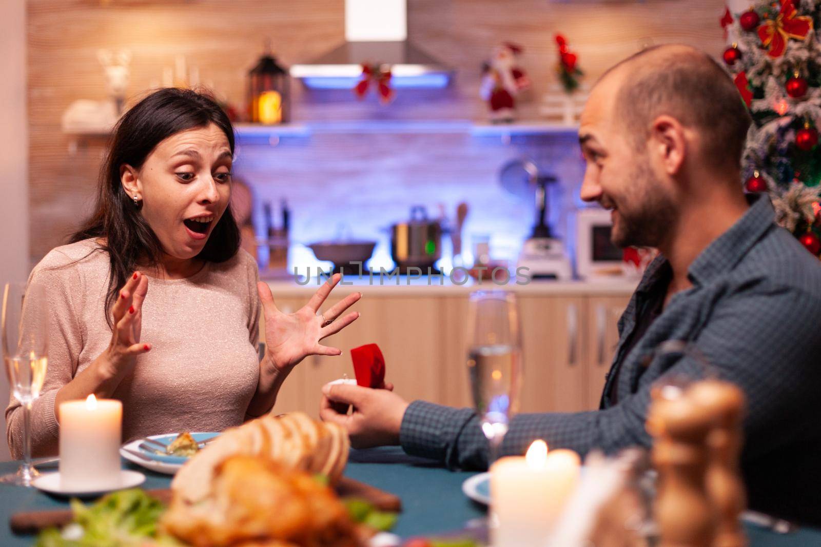Haapy man proposing girlfriend to marry him using diamond luxury expensive engagement ring celebrating christmas holiday enjoying winter season together. Cheerful couple sitting at xmas dinning table