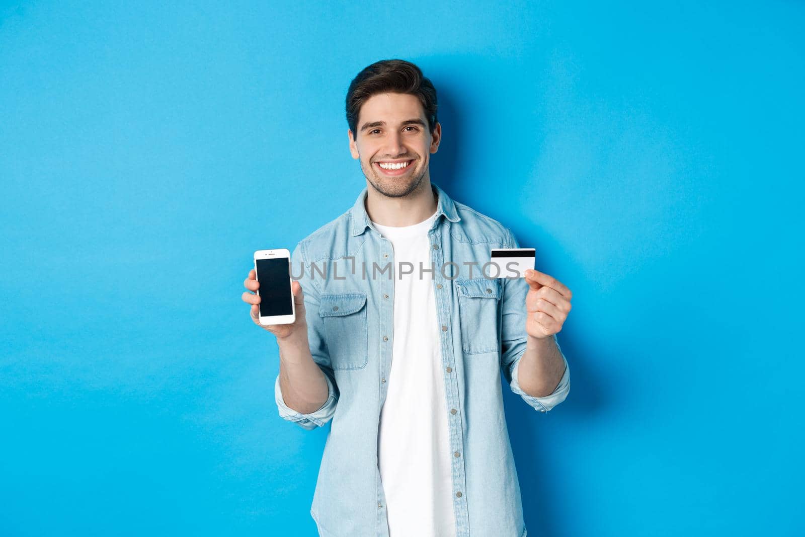 Young smiling man showing smartphone screen and credit card, concept of online shopping or banking.