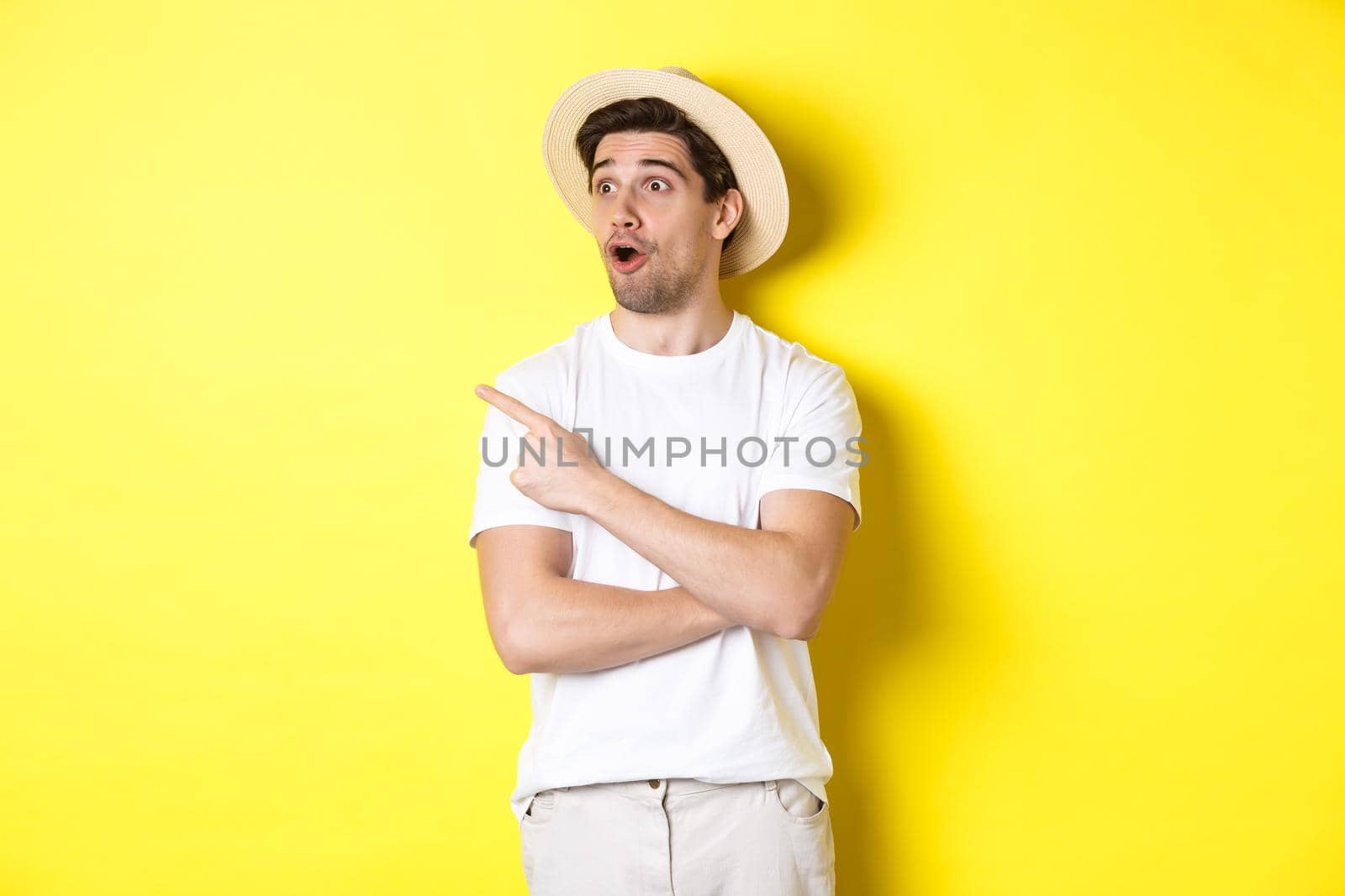 Concept of tourism and lifestyle. Excited handsome guy in straw hat checking out advertisement, pointing and looking at upper left corner logo, yellow background by Benzoix