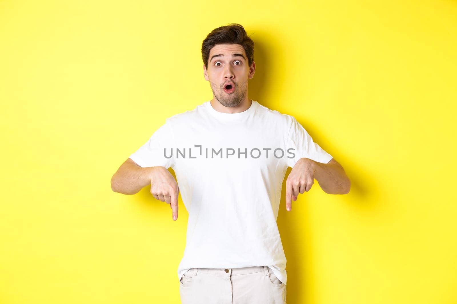 Image of impressed guy pointing fingers down, gasping amazed and looking at camera, standing over yellow background by Benzoix