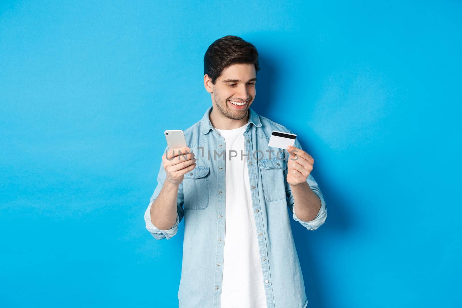 Handsome happy man paying for something online, holding credit card and mobile phone, purchase in internet, standing over blue background by Benzoix