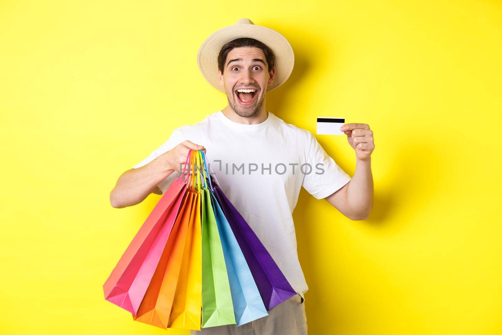 Happy guy going shopping on vacation, holding paper bags and showing credit card, standing over yellow background by Benzoix