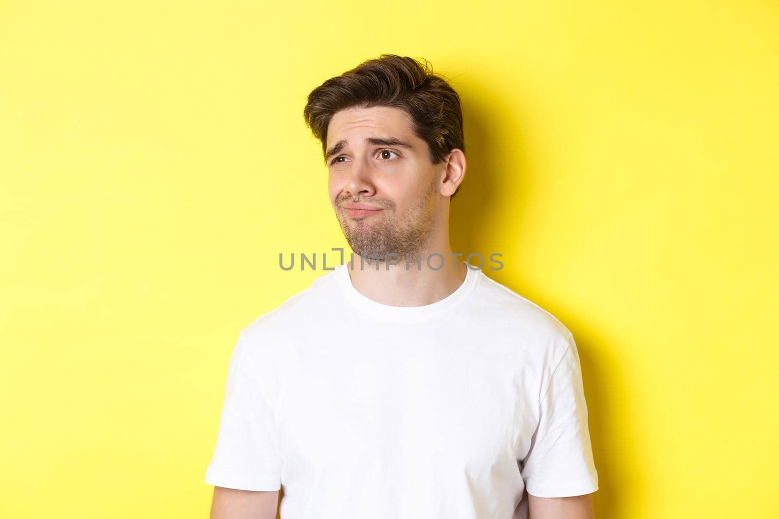 Reluctant guy in white t-shirt looking left, grimacing skeptical and displeased, standing over yellow background.
