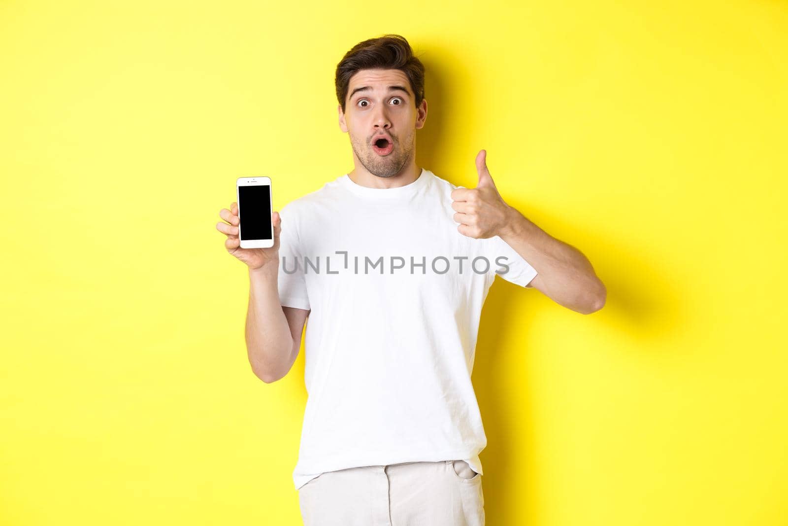 Impressed guy showing awesome smartphone app on screen and thumb-up, gasping amazed, standing against yellow background.