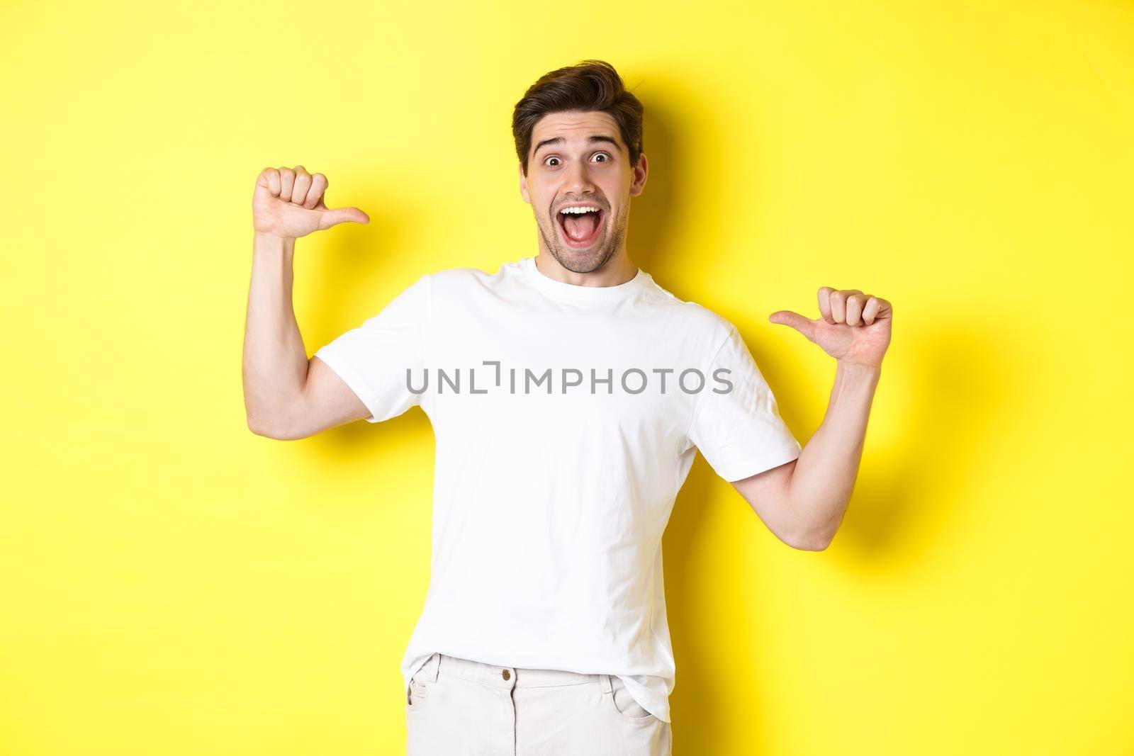 Excited man looking happy, pointing at himself with amazement, standing over yellow background.