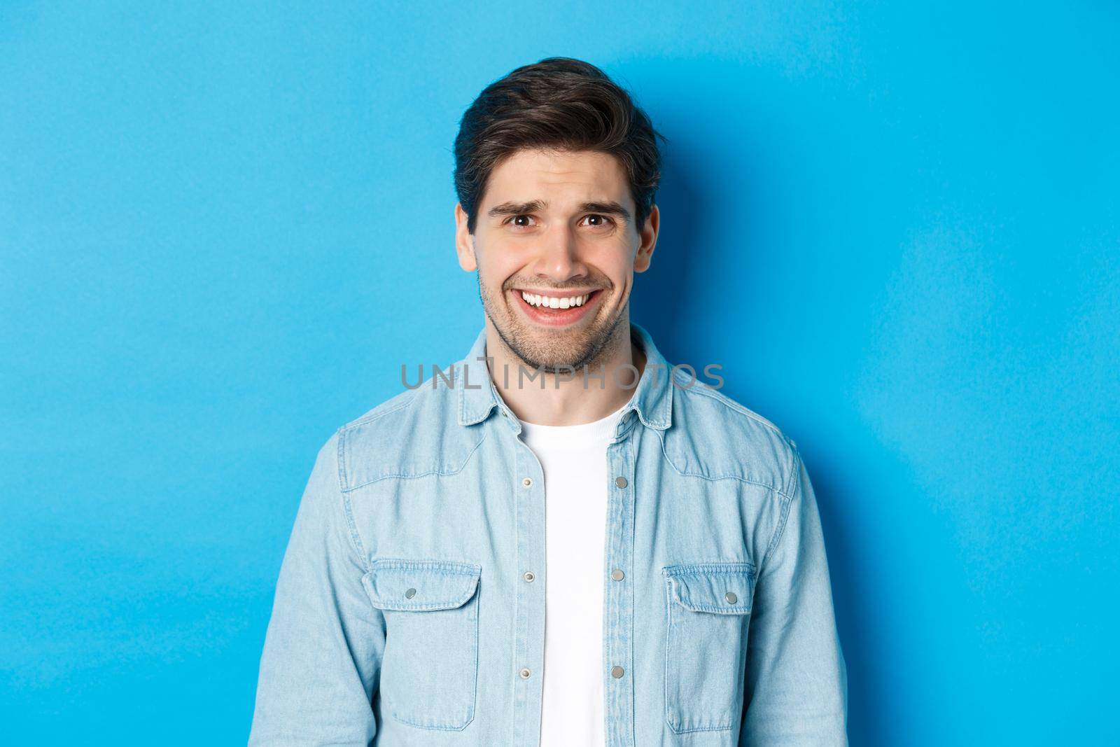 Close-up of young man feeling awkward, smile and cringe from uncomfortable situation, standing over blue background.