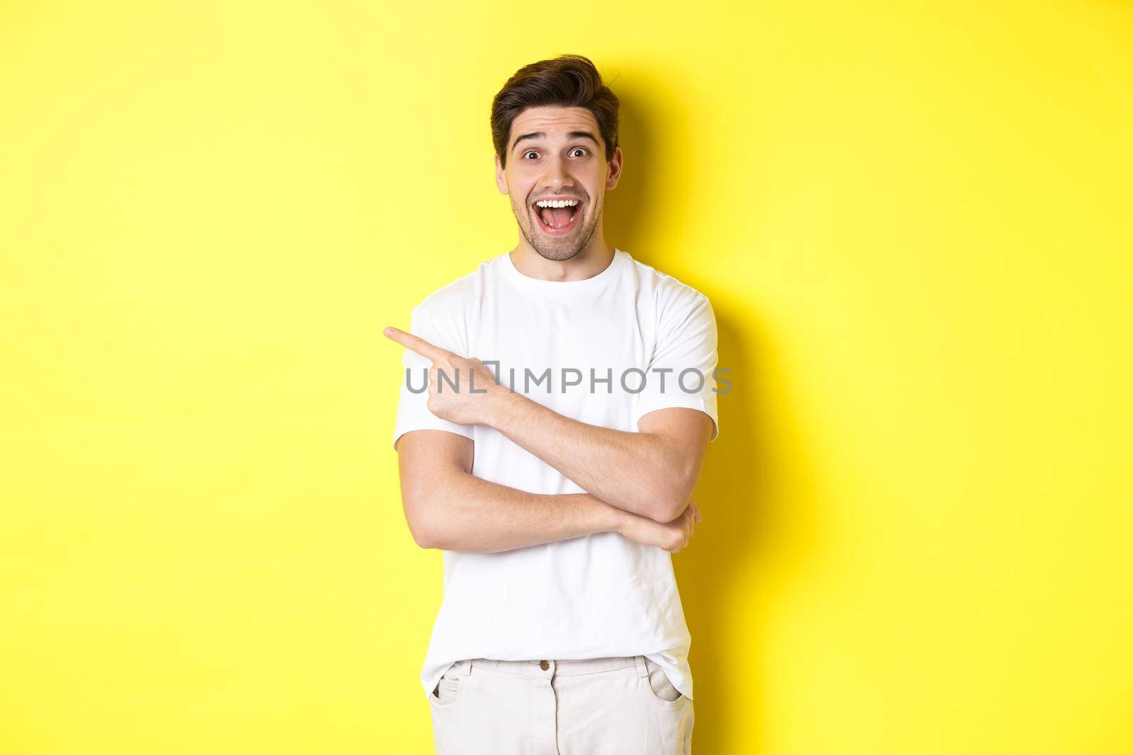 Happy man pointing finger left, showing advertisement on copy space, smiling amused, standing in white clothes against yellow background by Benzoix