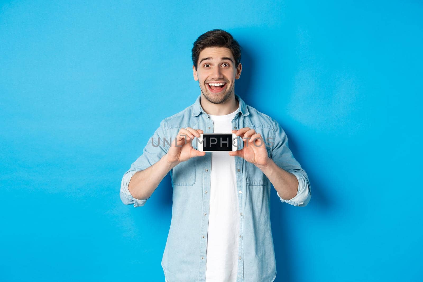 Amazed smiling man showing smartphone screen, internet promo offer, standing against blue background.