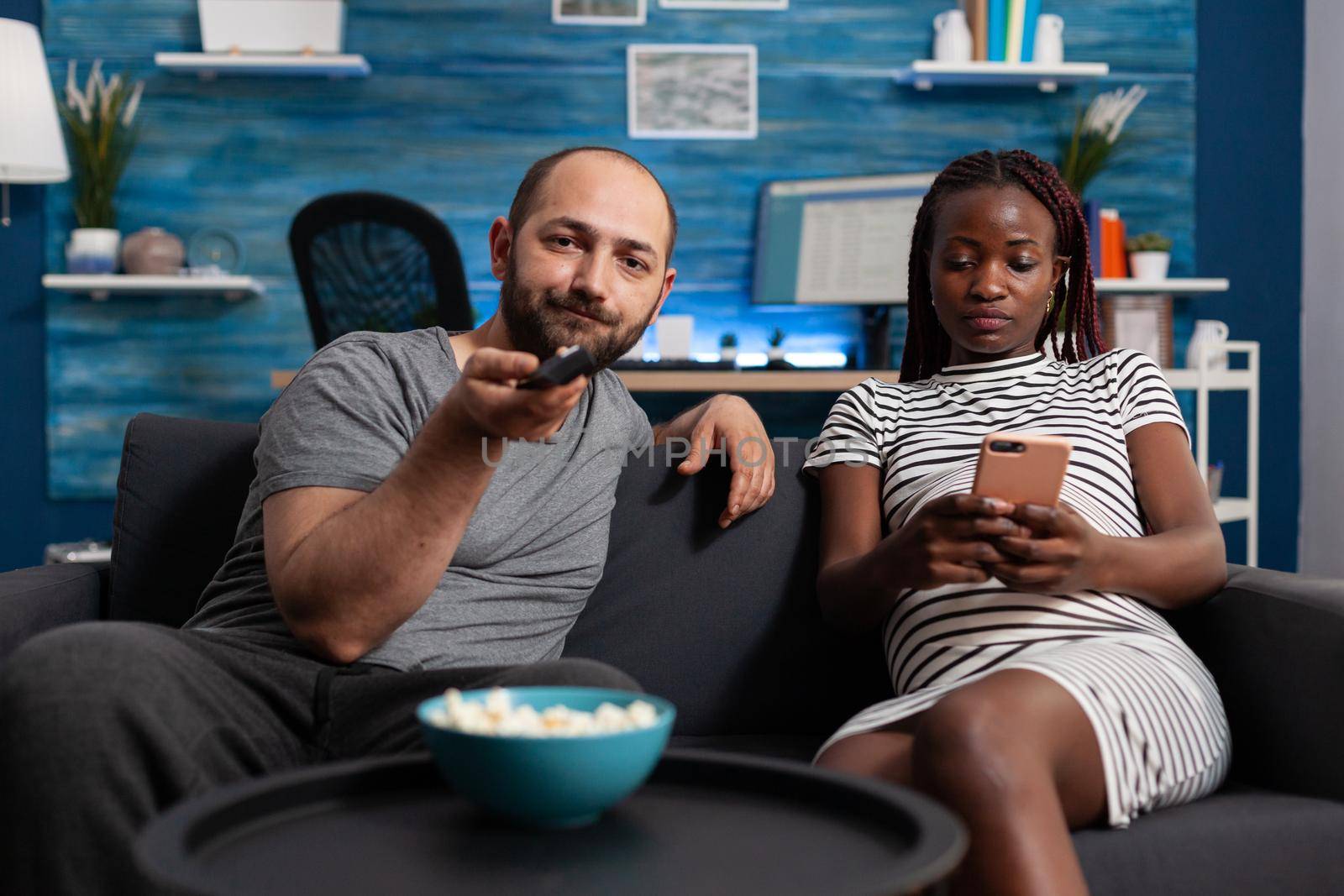 POV of interracial couple with technology expecting child at home. Caucasian husband watching television with TV remote control while african american pregnant wife looking at smartphone.