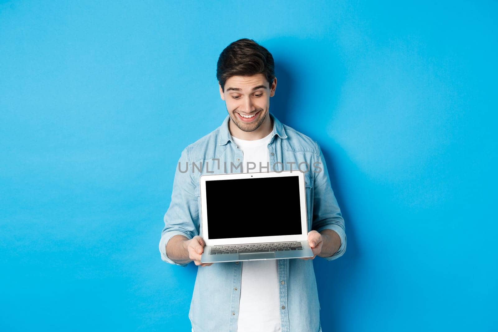 Excited handsome man looking at screen, showing promo offer on computer screen, smiling amazed, standing over blue background.
