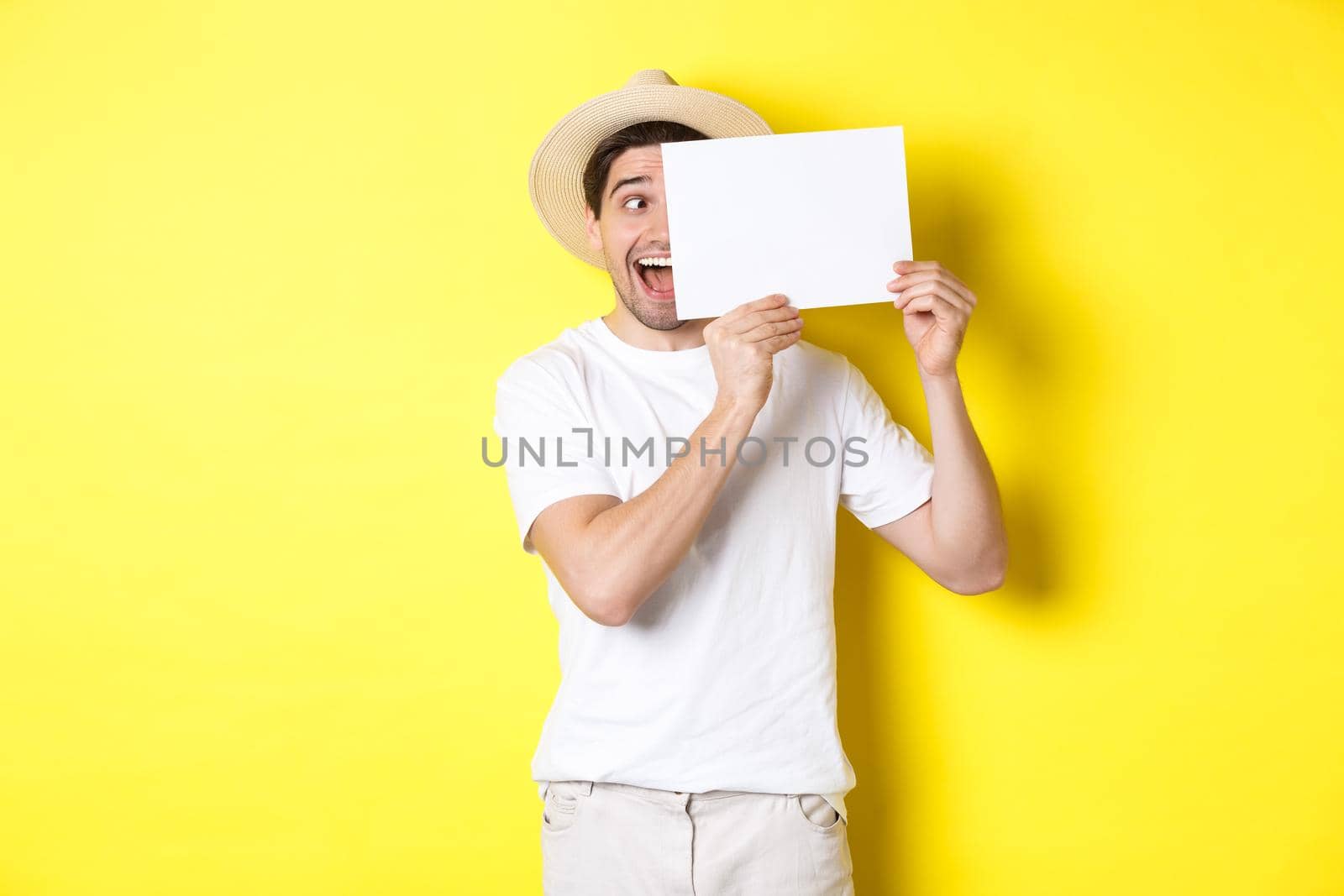 Excited man on vacation showing blank piece of paper for your logo, holding sign near face and smiling, standing against yellow background.