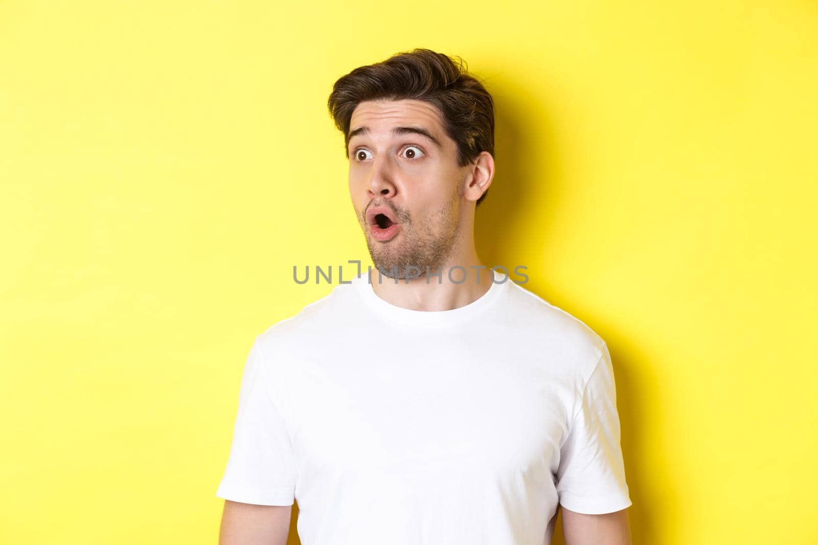 Close-up of impressed man looking left, gasping amazed, standing in white t-shirt against yellow background by Benzoix