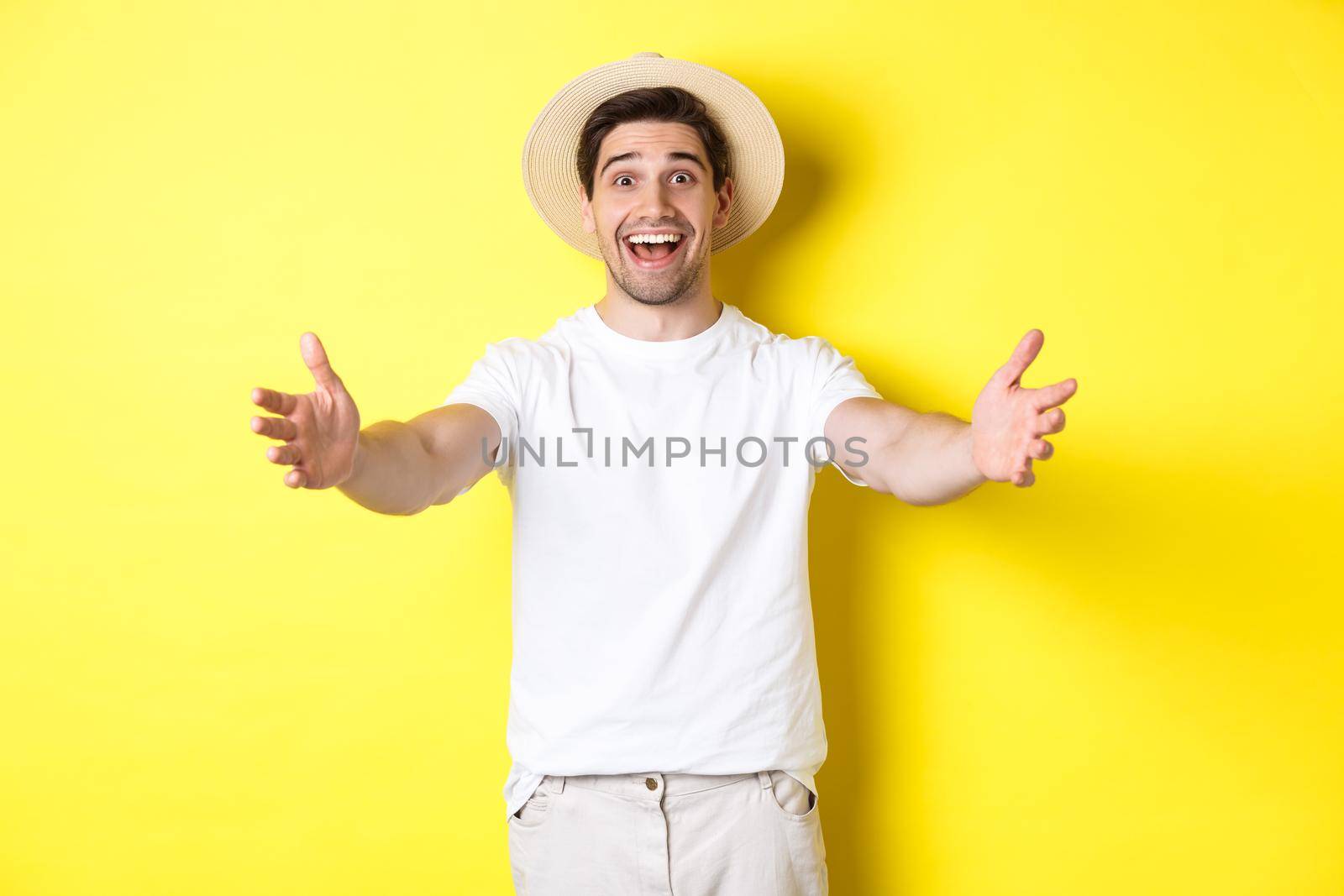 Concept of tourism and summer. Happy and friendly man traveller reaching hands for hug, greeting or welcome you, standing over yellow background.