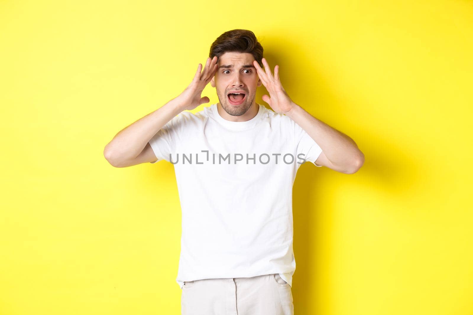 Frustrated guy looking shocked, panicking and holding hands on head, standing against yellow background.