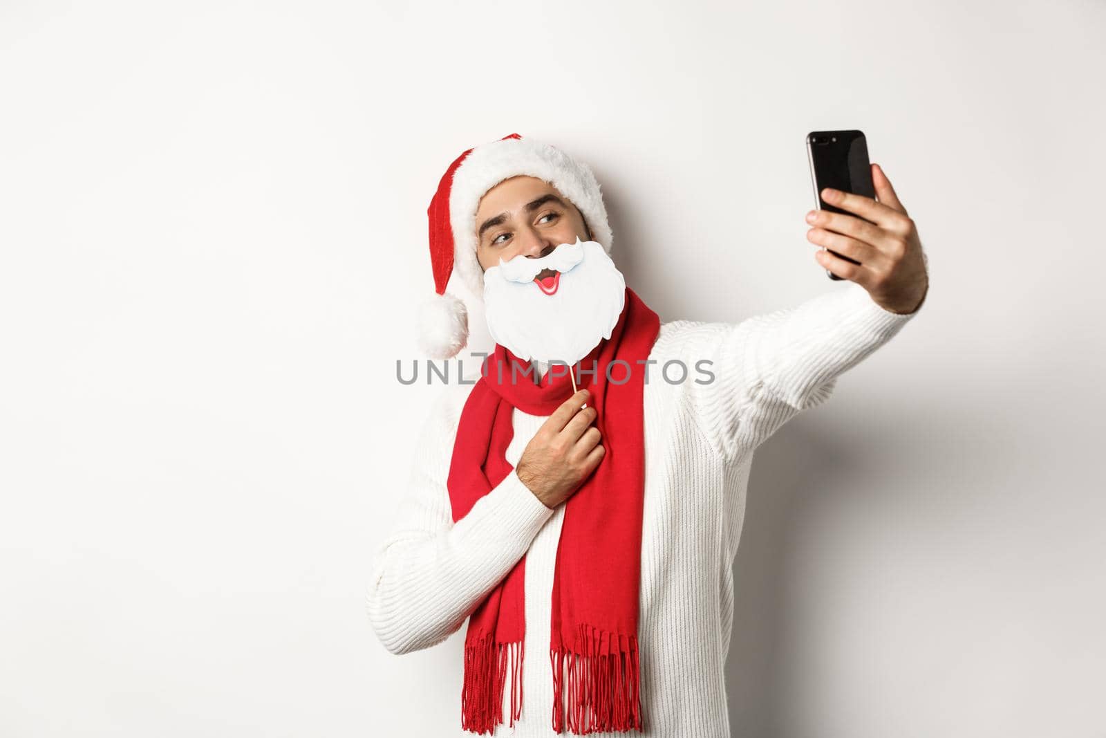 Christmas party and celebration concept. Young man taking selfie with funny white beard Santa mask and hat, posing for photo on mobile phone, studio background.