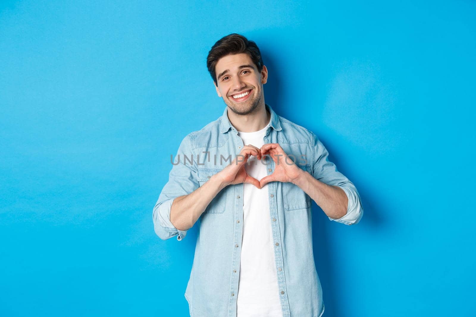 Handsome man smiling, showing heart gesture and looking at camera, saying I love you, standing against blue background.