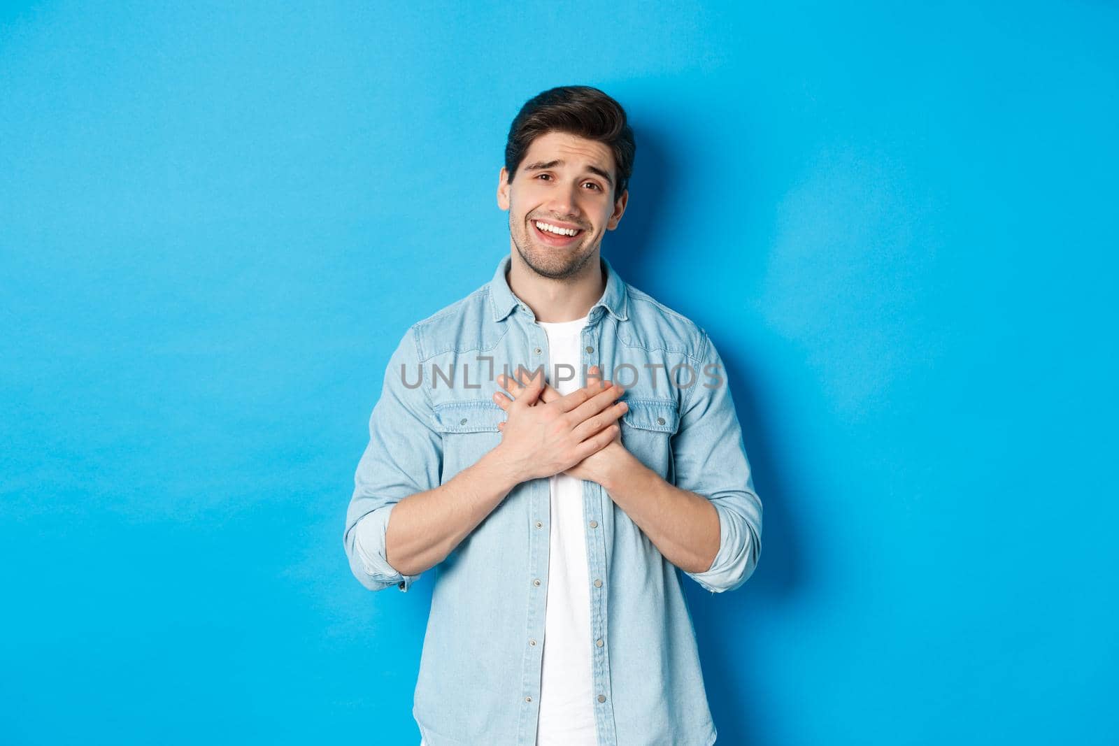 Portrait of flattered handsome man thanking you, holding hands on heart and smiling, looking grateful, standing over blue background.