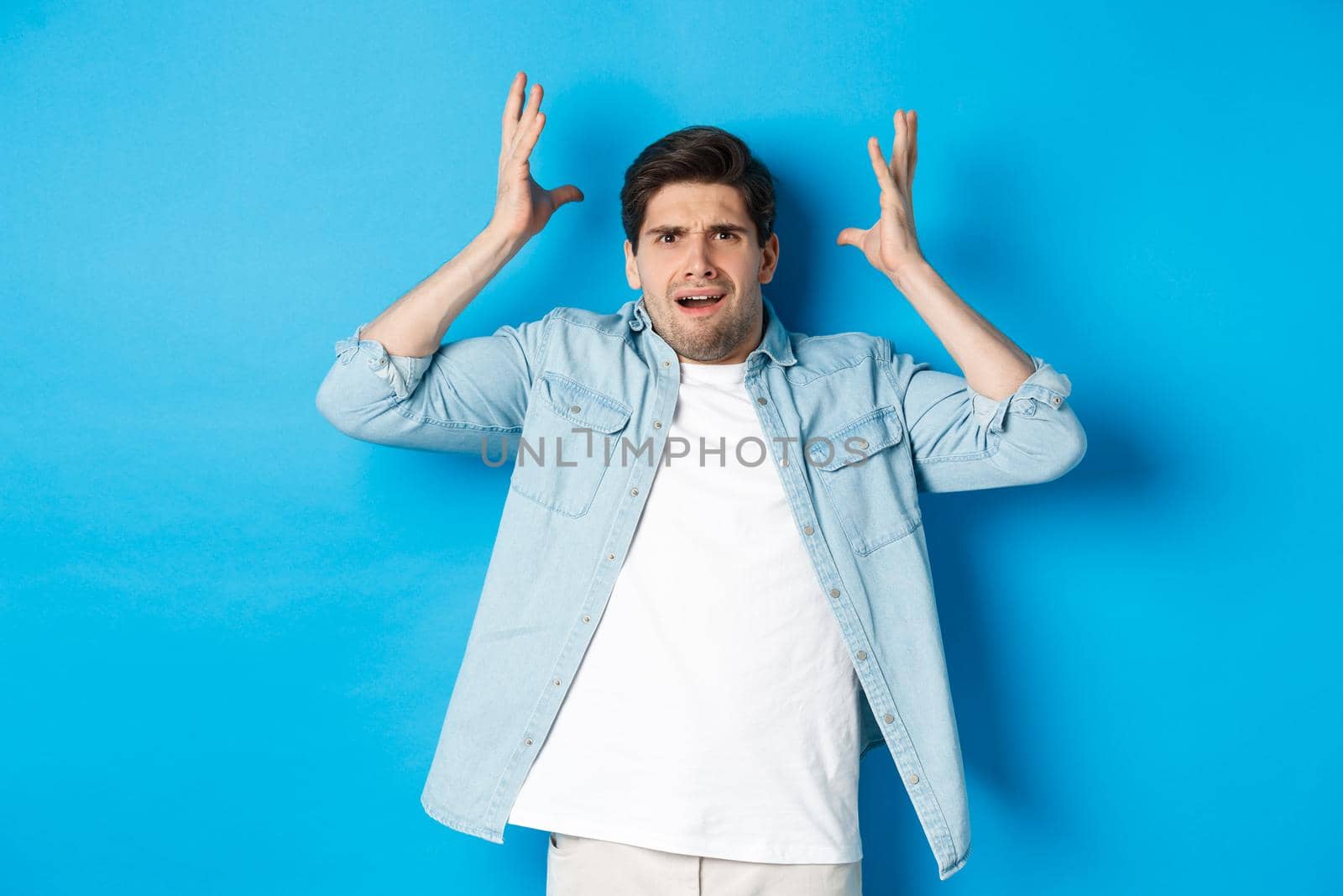 Confused and bothered man holding hands near head, looking frustrated, standing upset against blue background.