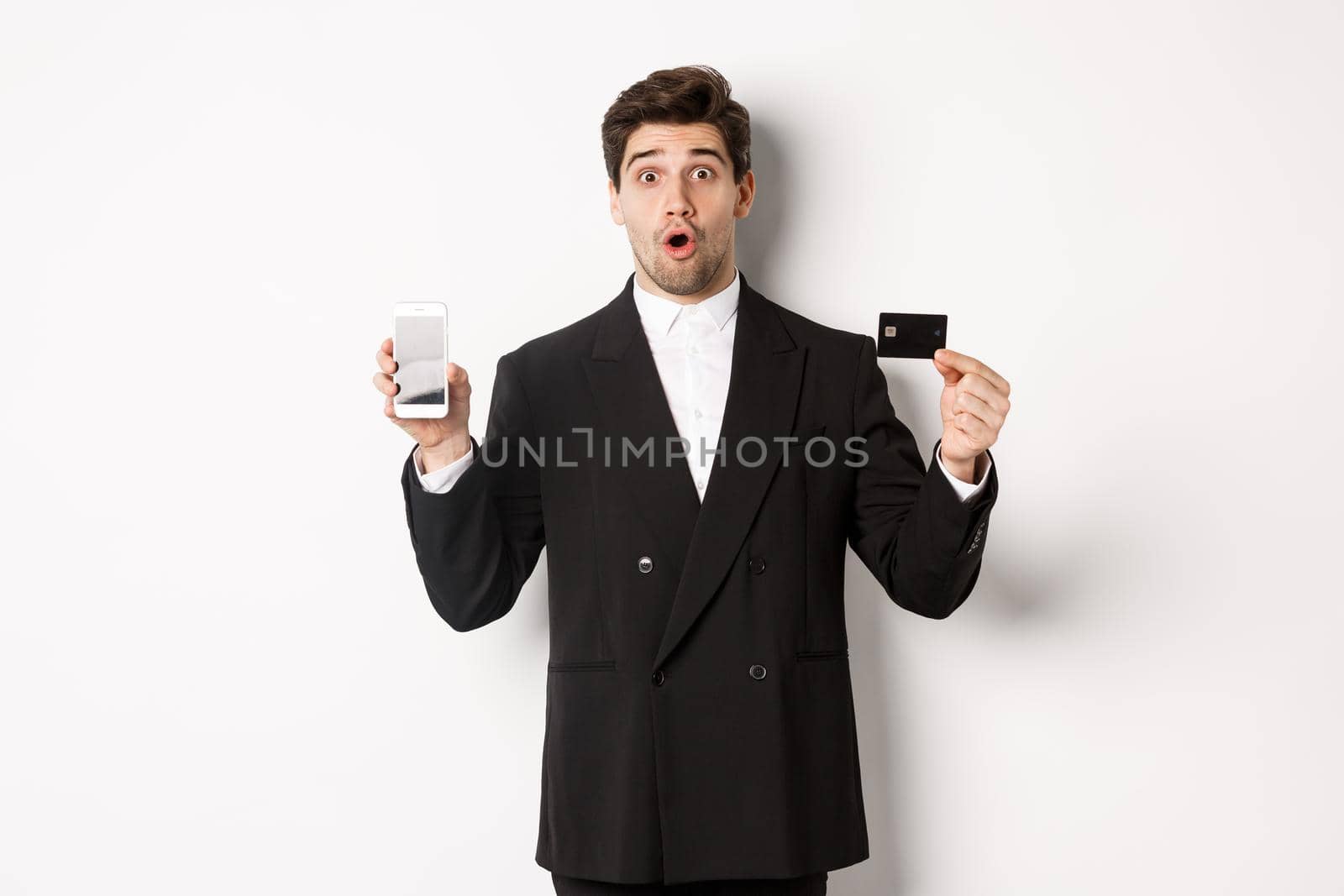 Image of handsome businessman in black suit, looking amazed and showing credit card with mobile phone screen, standing against white background.