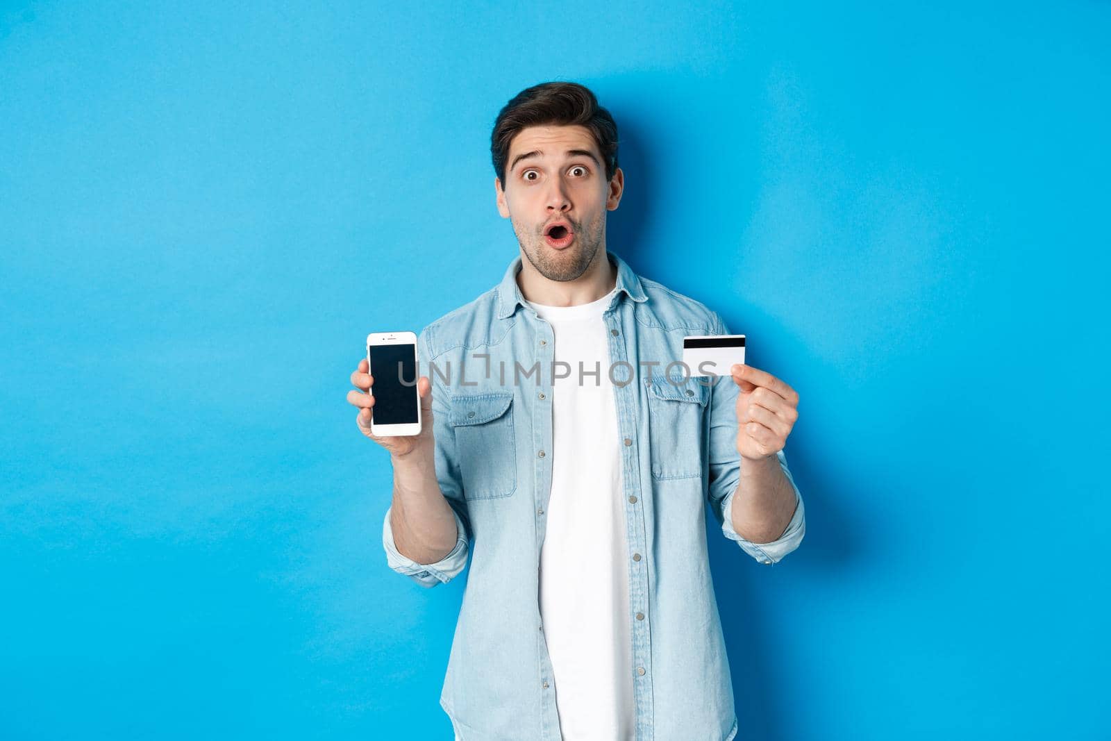 Amazed young man showing mobile cell phone screen and credit card, shop online, standing against blue background.