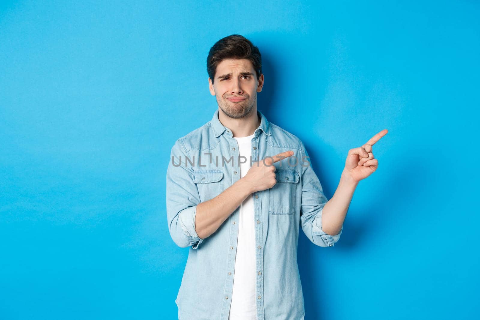 Portrait of skeptical adult guy pointing fingers right and smirking, exress disappointment and doubt, standing against blue background.