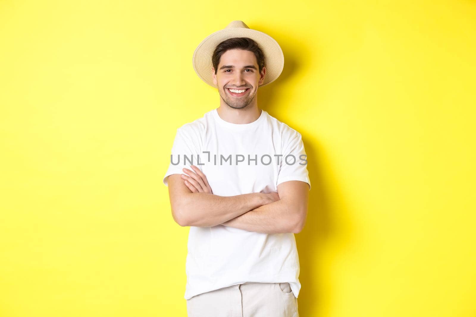 Tourism. Handsome young man looking happy, wearing straw hat for travelling, cross hands on chest and smiling, standing over yellow background.