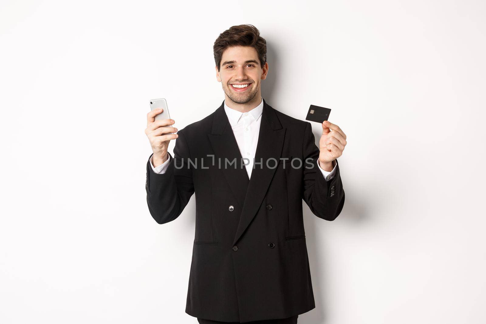 Handsome businessman in black suit smiling, showing credit card and money, standing against white background by Benzoix